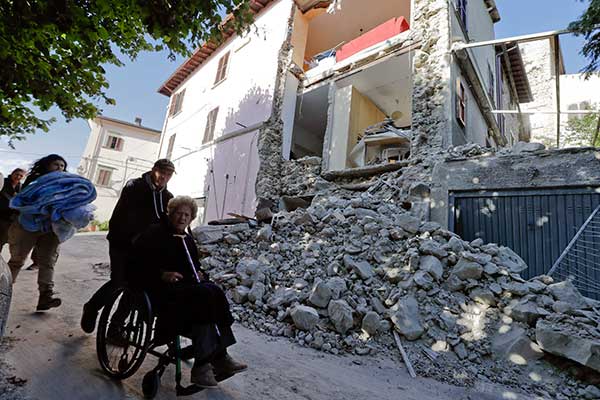A woman on a wheelchair is pushed past a partially collapsed building is seen in the background following an earthquake, in Accumoli.