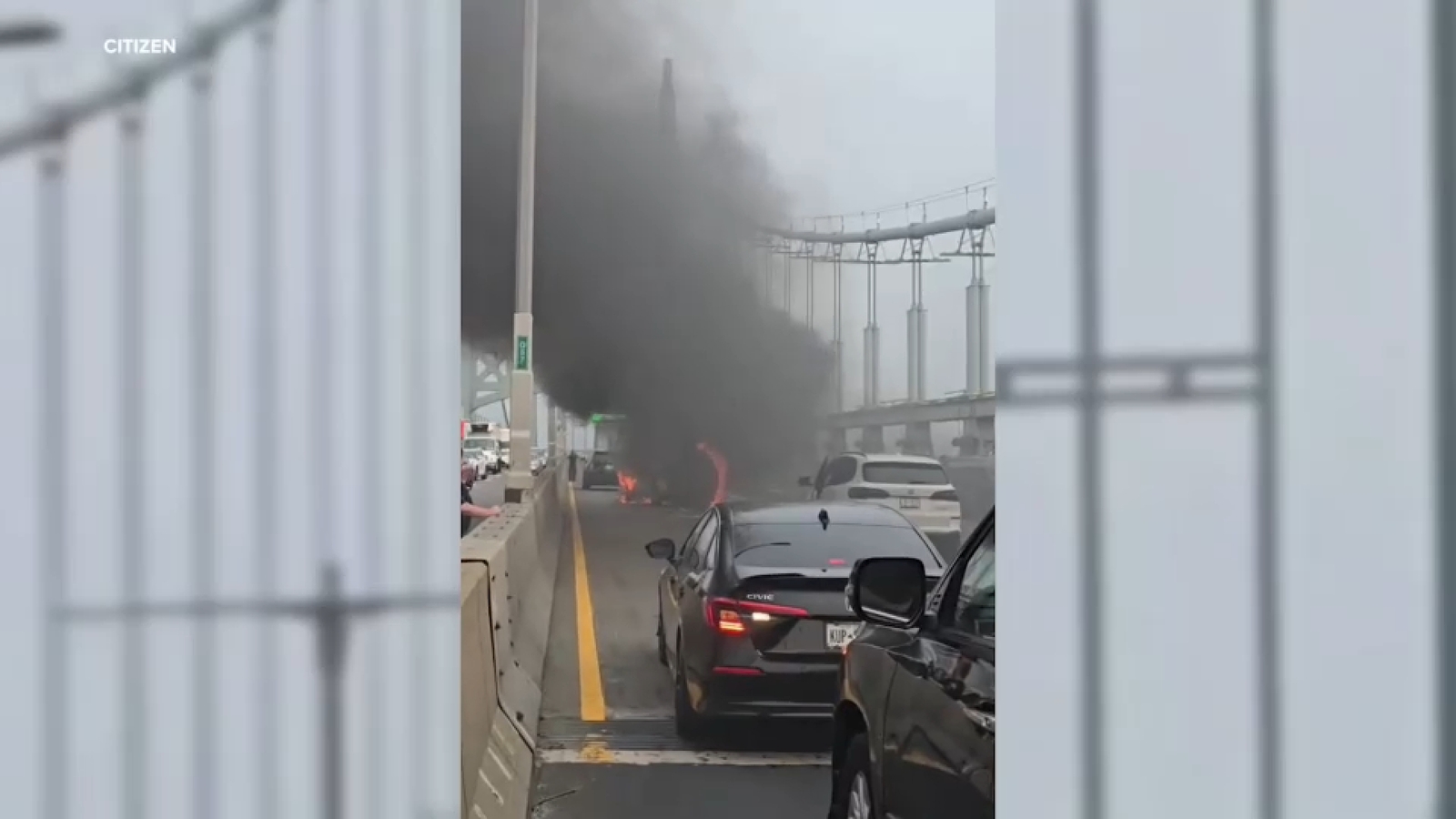 Multiple cars stopped on a bridge lane with a vehicle on fire ahead producing large black smoke that fills the roadway; other cars and concrete barriers line the bridge.