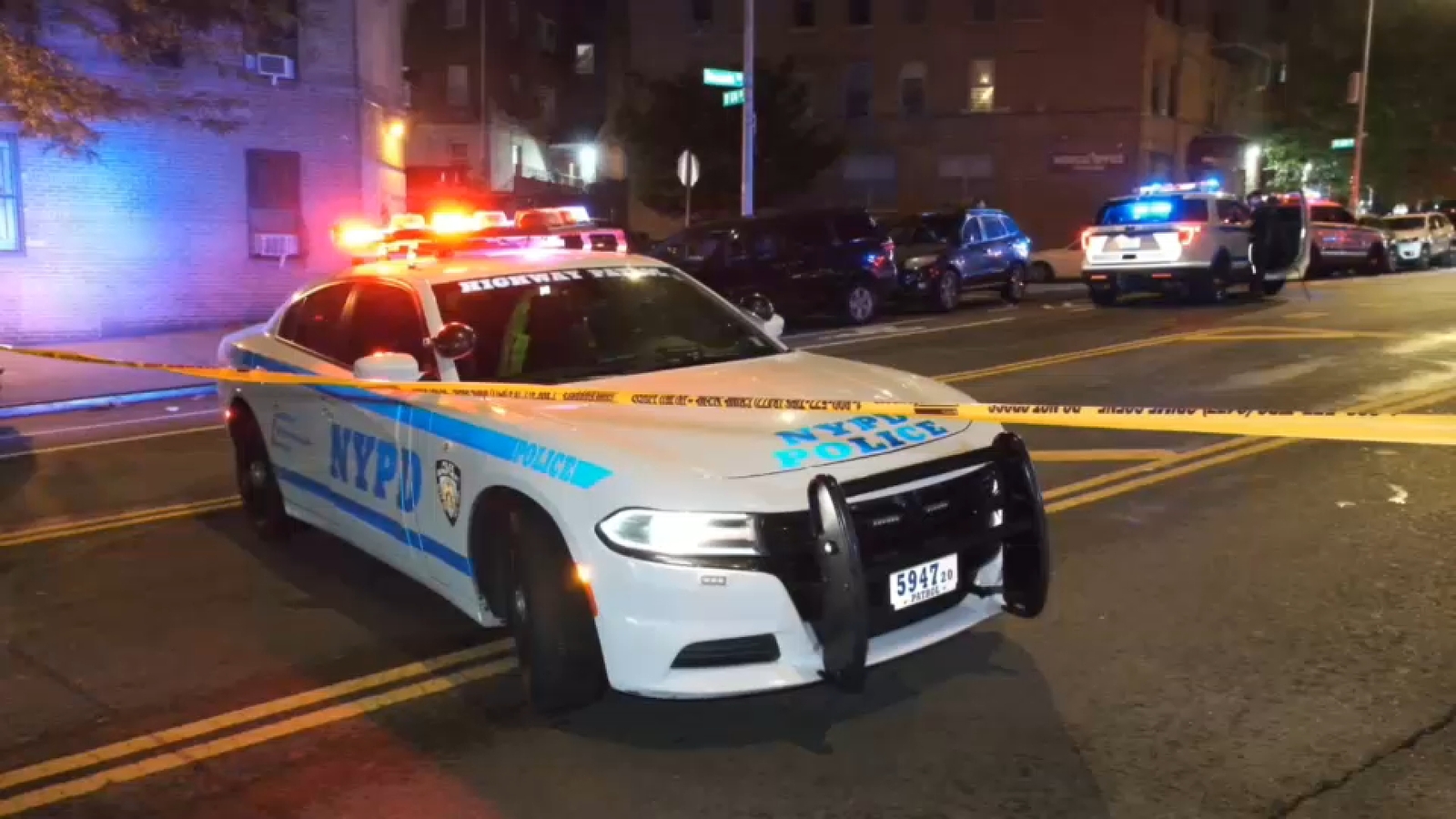 White NYPD patrol car with flashing red and blue lights parked diagonally across a city street at night. Yellow police tape stretches across the foreground and several other police vehicles and parked cars are visible along the curb in front of apartment buildings.