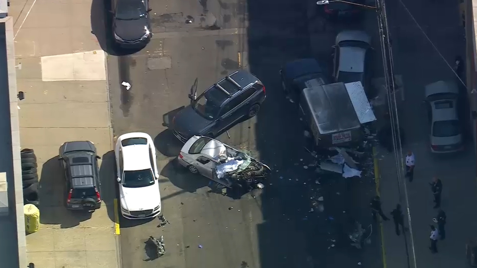 Aerial photo of a street crash in Astoria: a heavily damaged silver sedan with its front crumpled sits near a damaged box truck and several other vehicles. Debris is scattered across the roadway and several people, including emergency responders, stand on the sidewalk to the right.