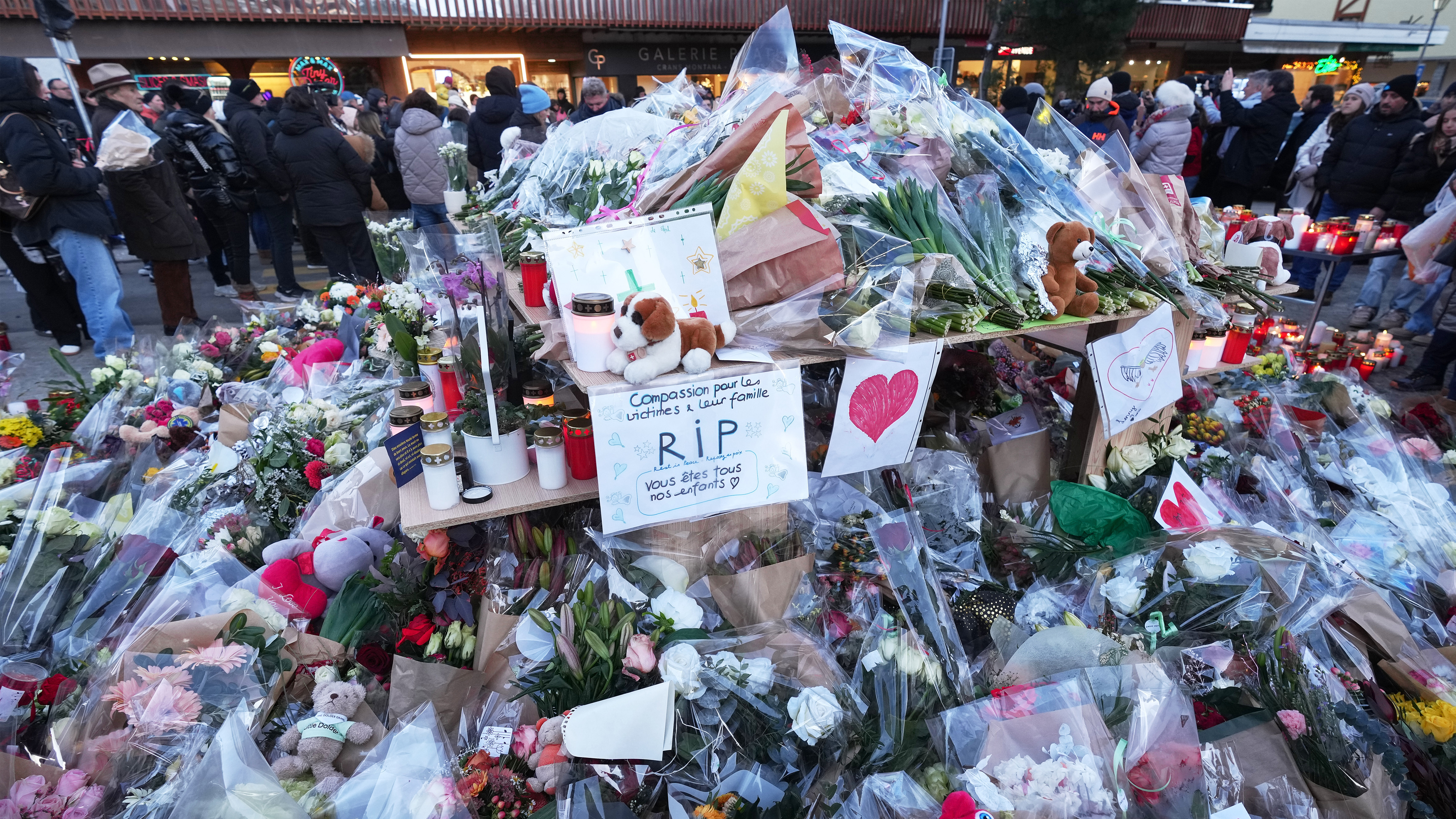 People stand around floral tributes and candles placed outside the sealed off Le Constellation bar in Crans-Montana, Swiss Alps, Switzerland, Saturday, Jan. 3, 2026.
