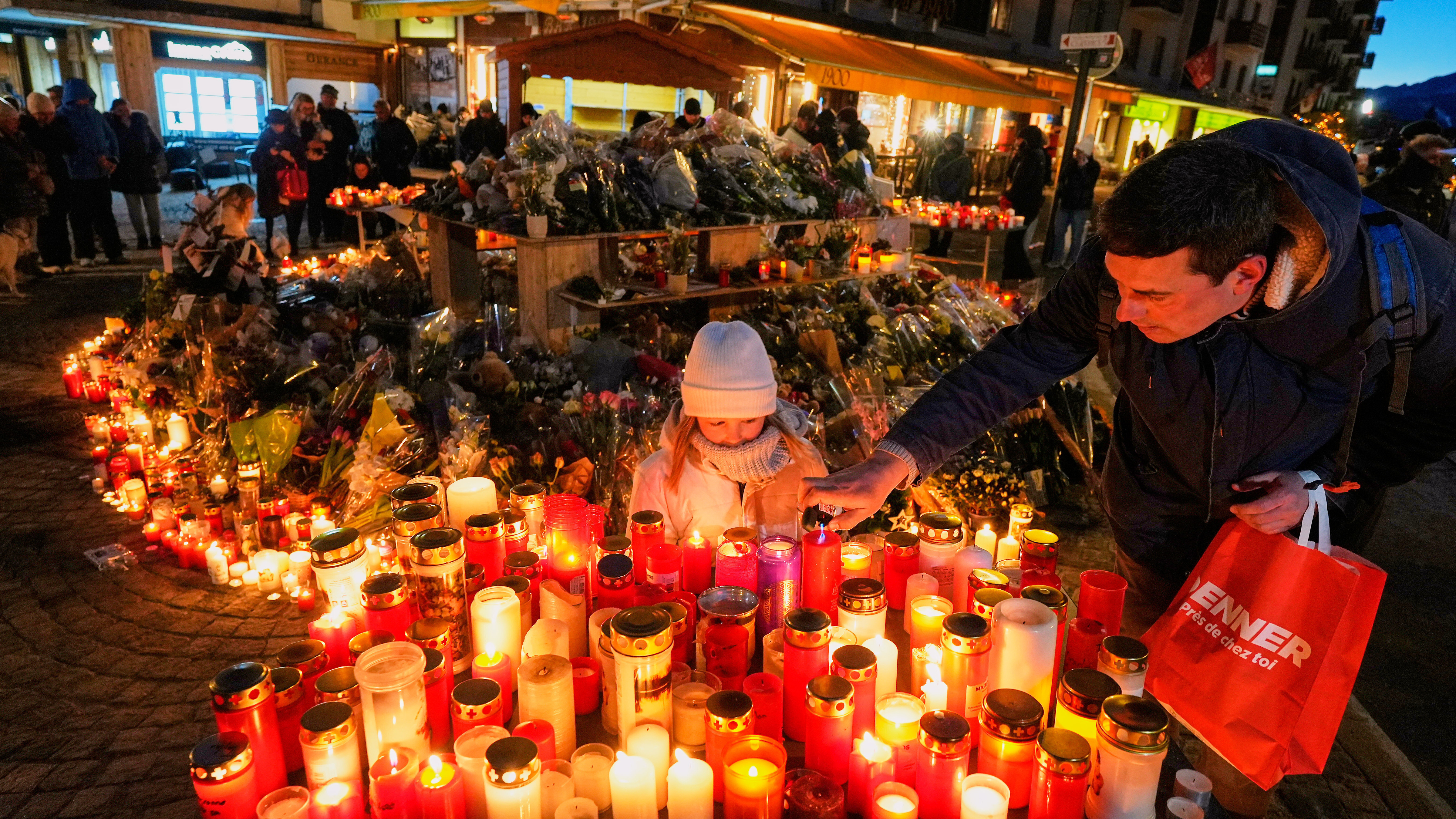 A child watches as an adult places a candle outside the sealed off Le Constellation bar in Crans-Montana, Swiss Alps, Switzerland, Saturday, Jan. 3, 2026, where a devastating fire left dead and injured during the New Year's celebrations. 