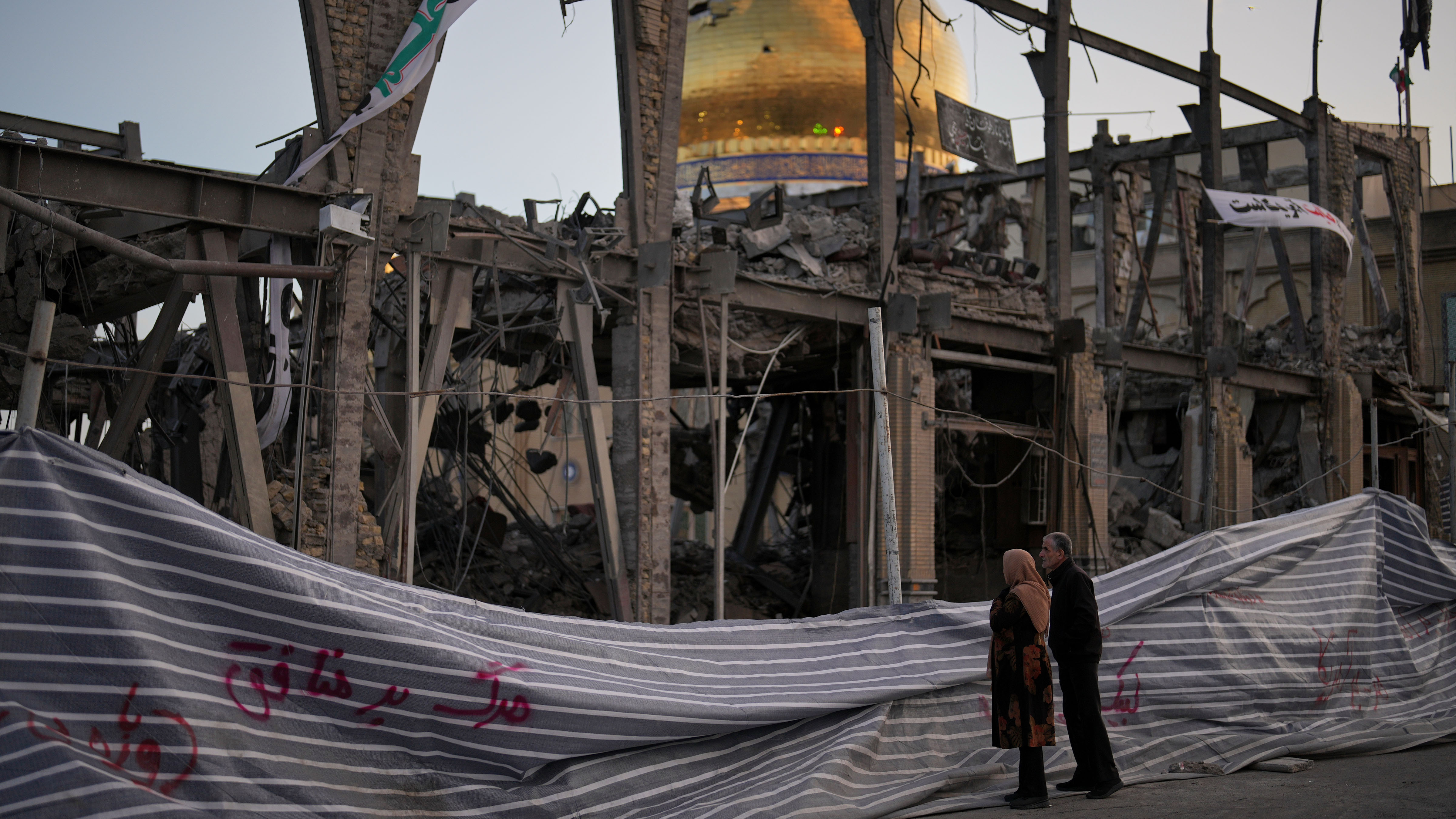 Pedestrians look at a destroyed building within the Grand Hosseiniyeh, with the mosque visible in the background, which officials say was hit by U.S.-Israeli airstrikes Tuesday.