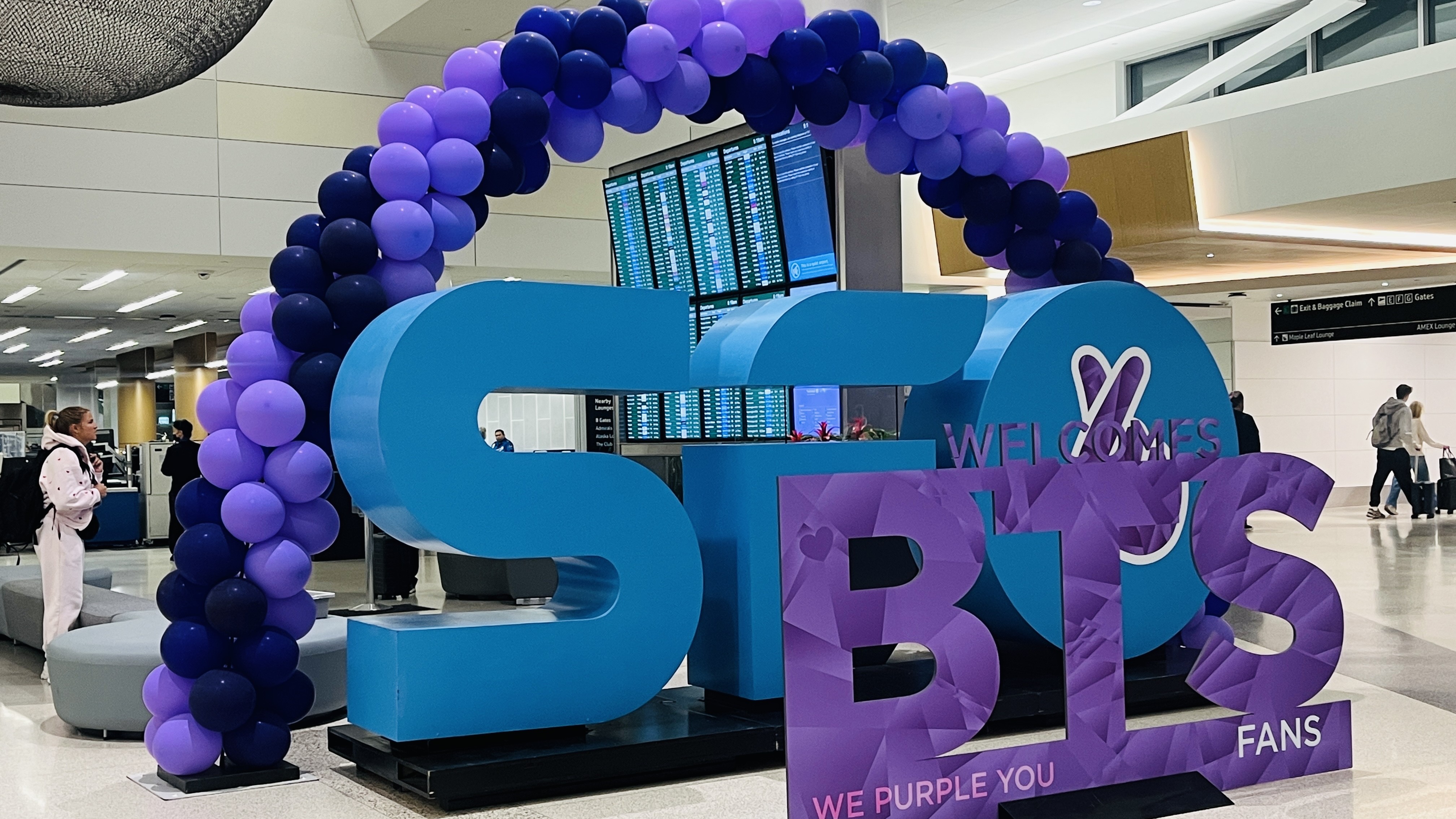 The image shows welcome signs and balloons at San Francisco International Airport ahead of Korean pop group BTS's arrival for their comeback tour series of concerts