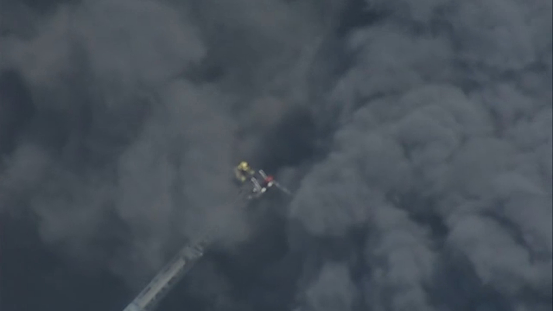 A firefighter is barely visible on a ladder amid thick black smoke from a massive auto shop fire in South Gate on Sunday, May 24, 2026.