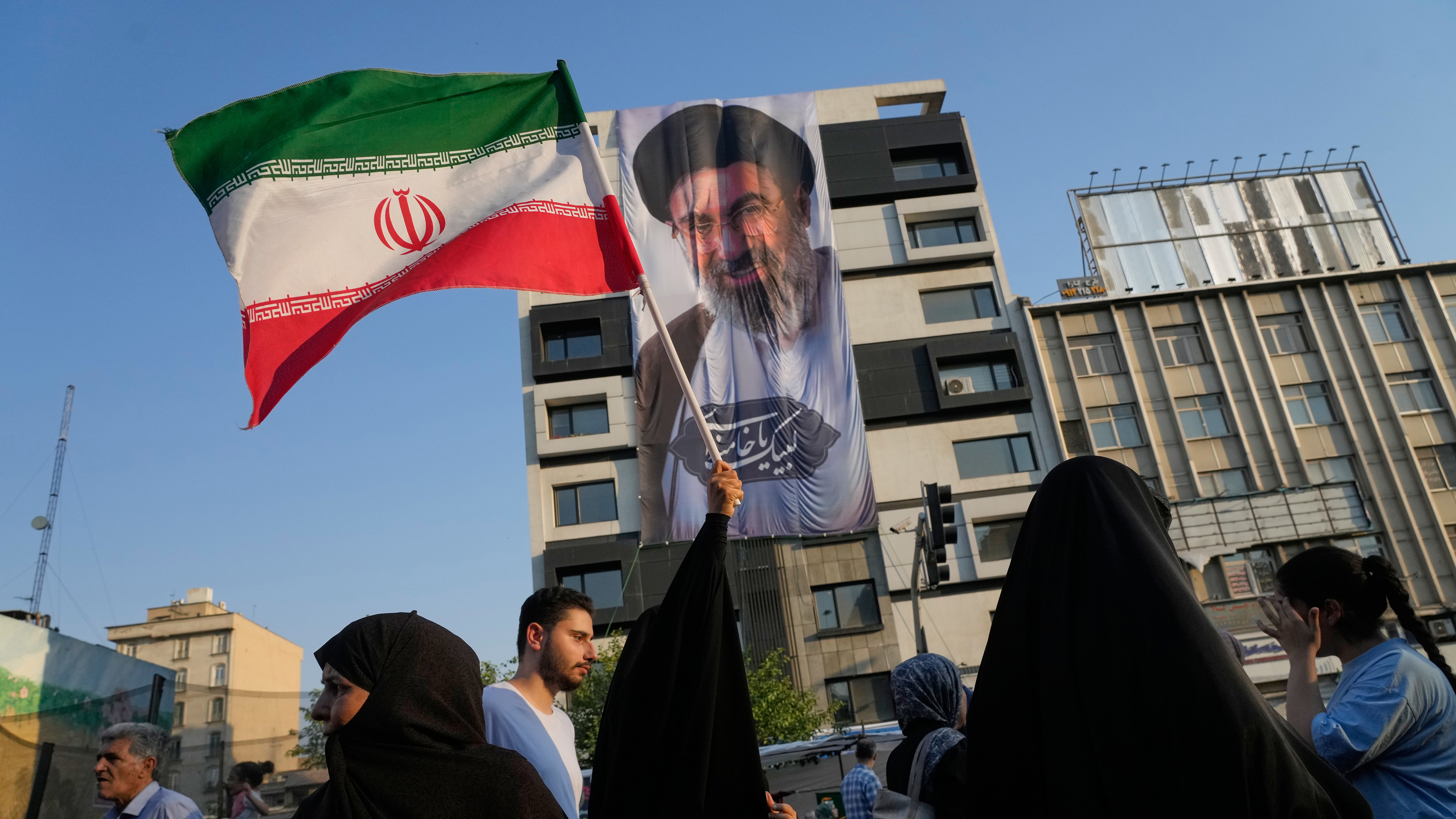 A woman holds up an Iranian flag in front of a banner showing a portrait of the Supreme Leader Ayatollah Mojtaba Khamenei during a street festival