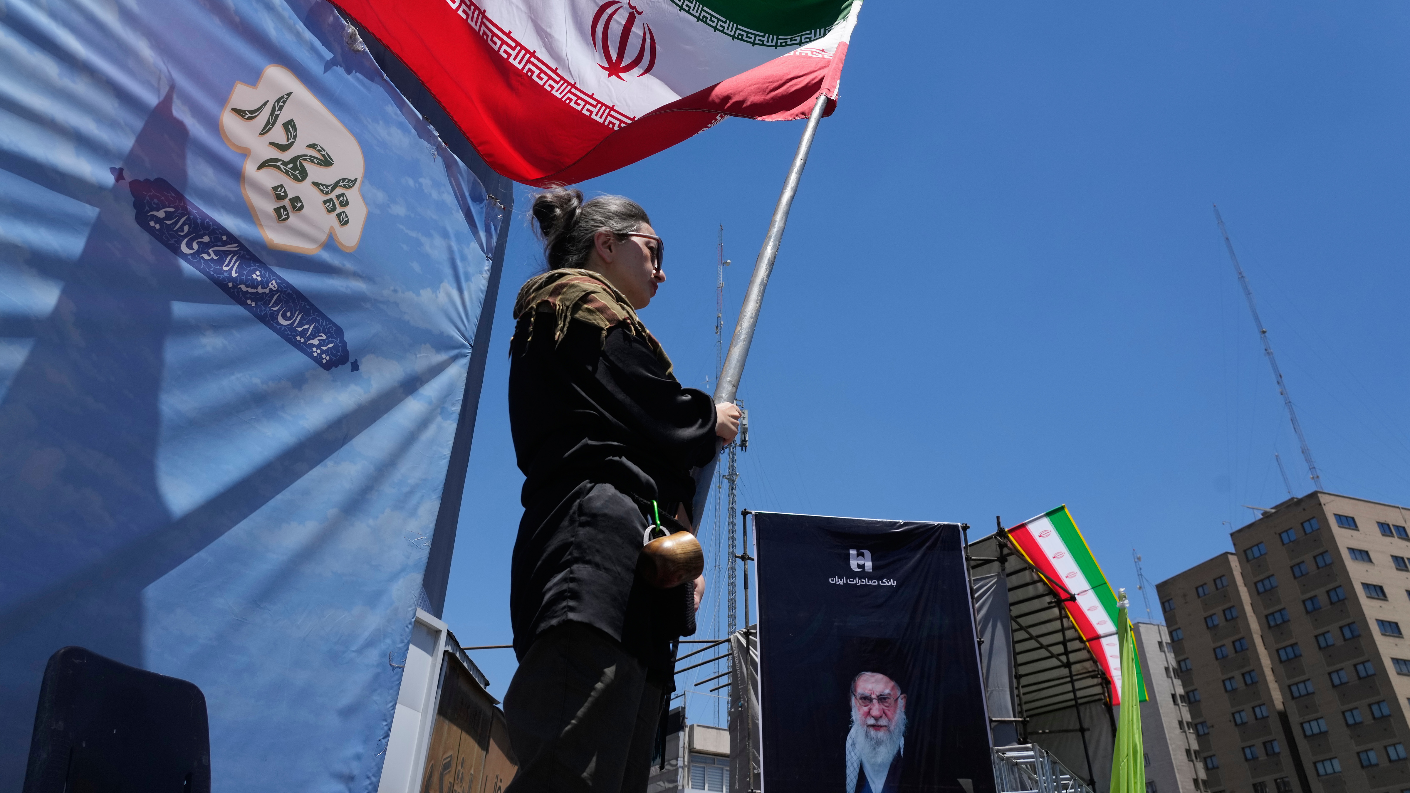 A woman waves an Iranian flag during a pro-government campaign as a portrait of the slain Supreme Leader Ayatollah Ali Khamenei, who was killed in U.S. and Israeli strikes on Feb. 28, is displayed at right, in downtown Tehran, Iran, Monday, June 15, 2026.